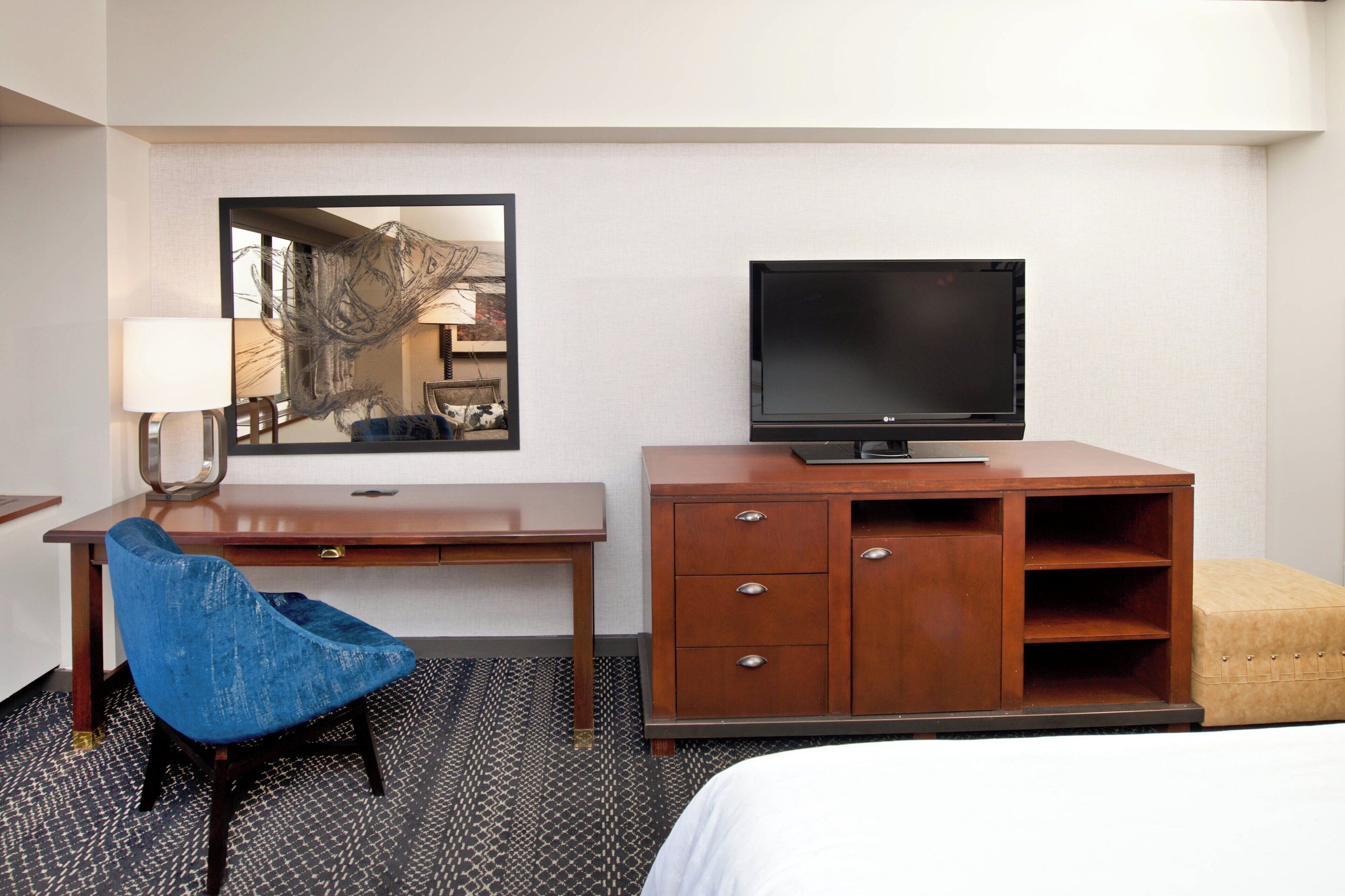Habitación de hotel con una cama, un escritorio de madera con televisor, una silla azul y un espejo que refleja la habitación.