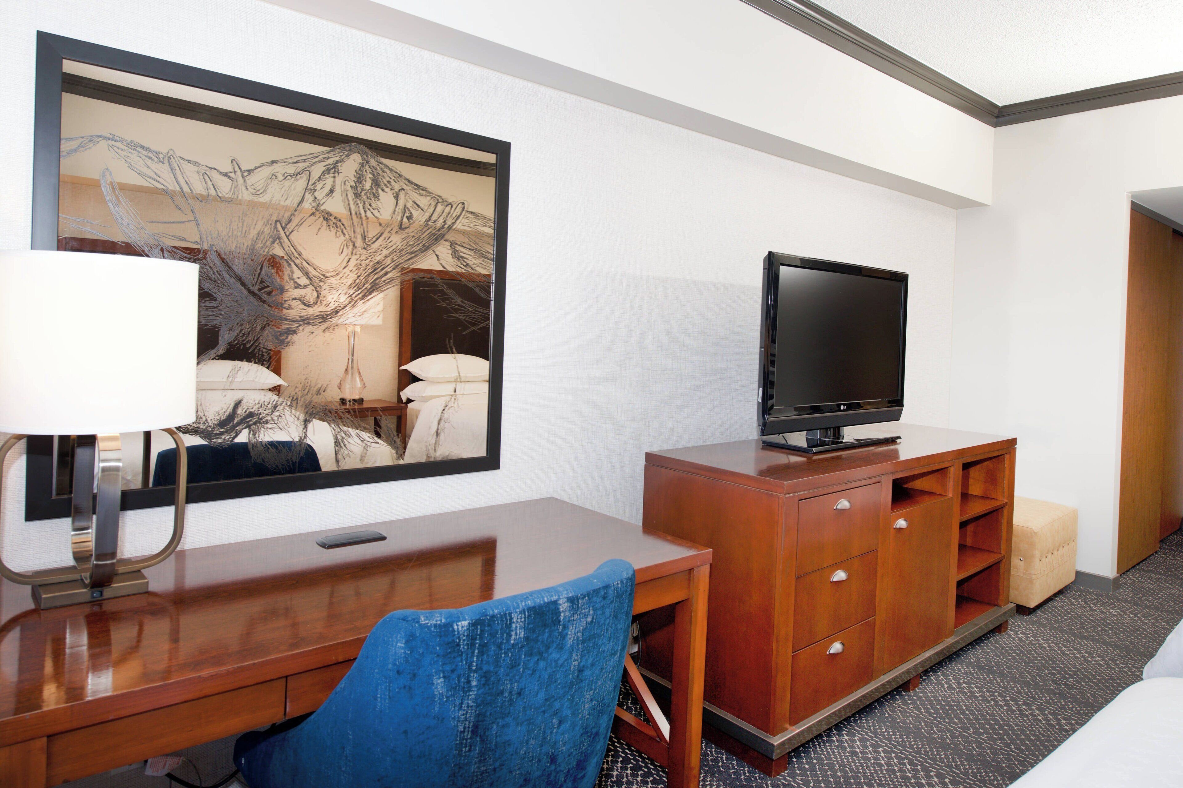 A hotel room with a wooden desk, a television on a wooden cabinet, a blue chair, a large framed artwork on the wall, and a bed with a white headboard.