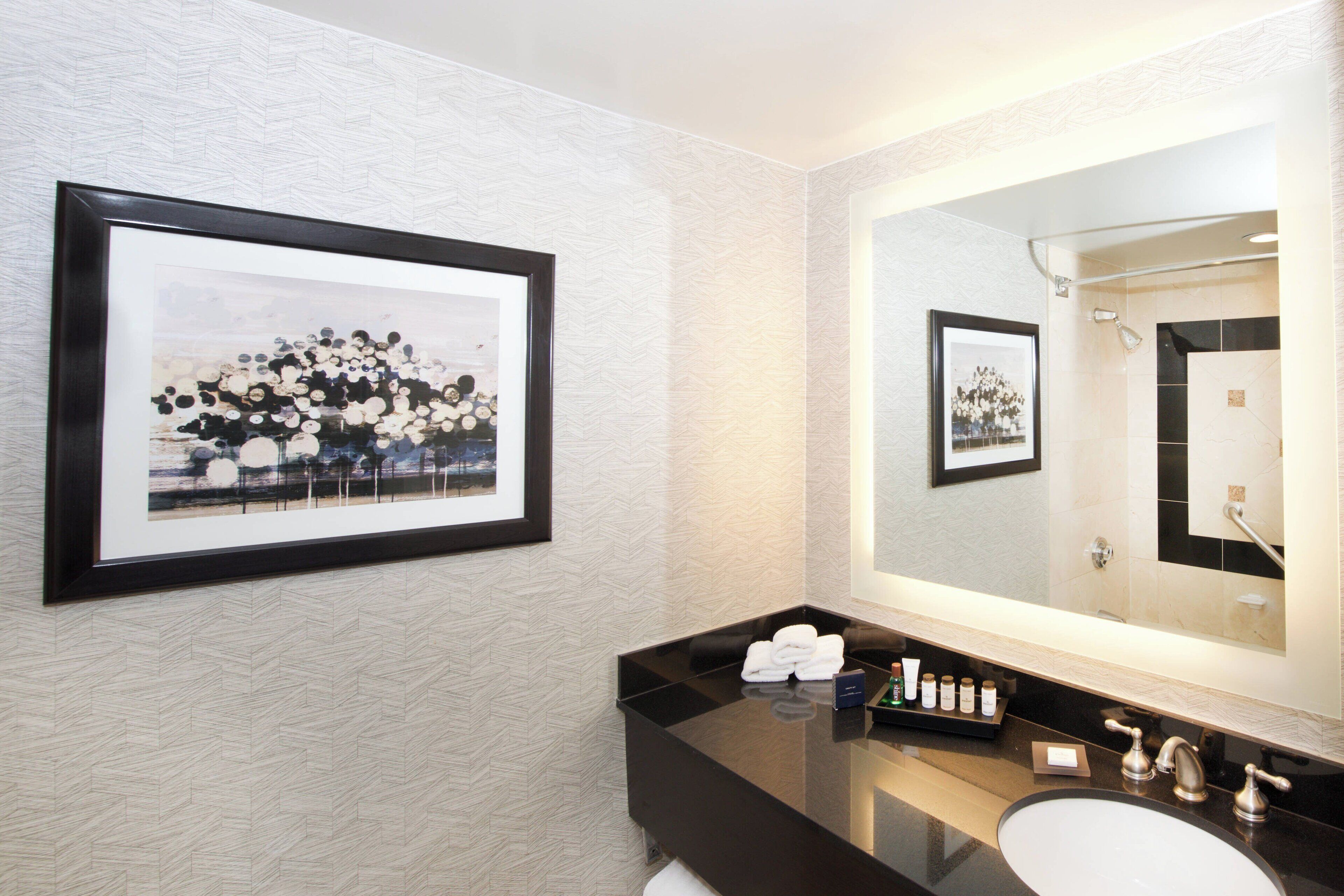 Un baño moderno con encimera negra, lavabo blanco y un espejo grande. Hay cuadros enmarcados en la pared y artículos de aseo en la encimera.