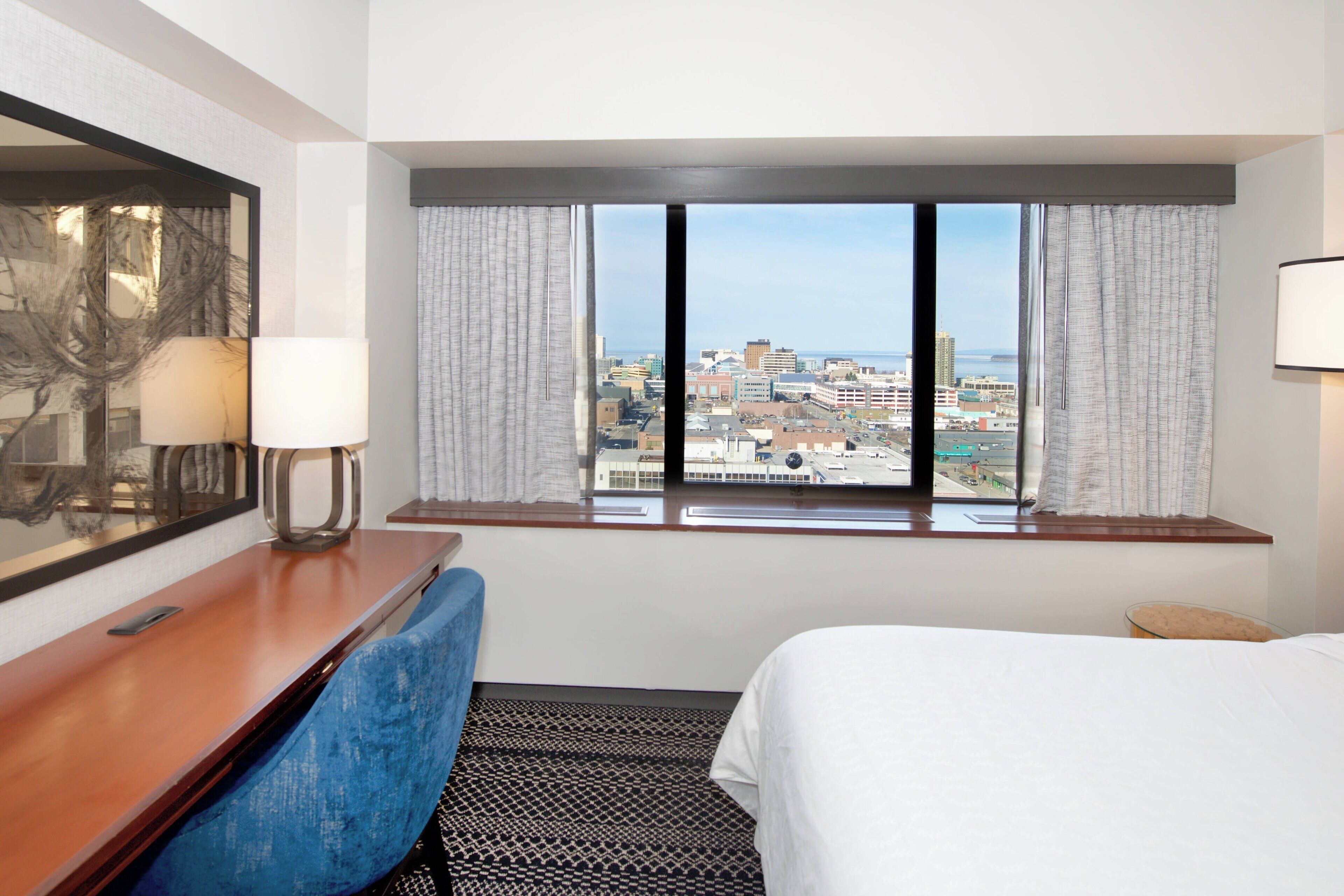 A hotel room with a large window, a bed, a desk with a lamp, and a chair.