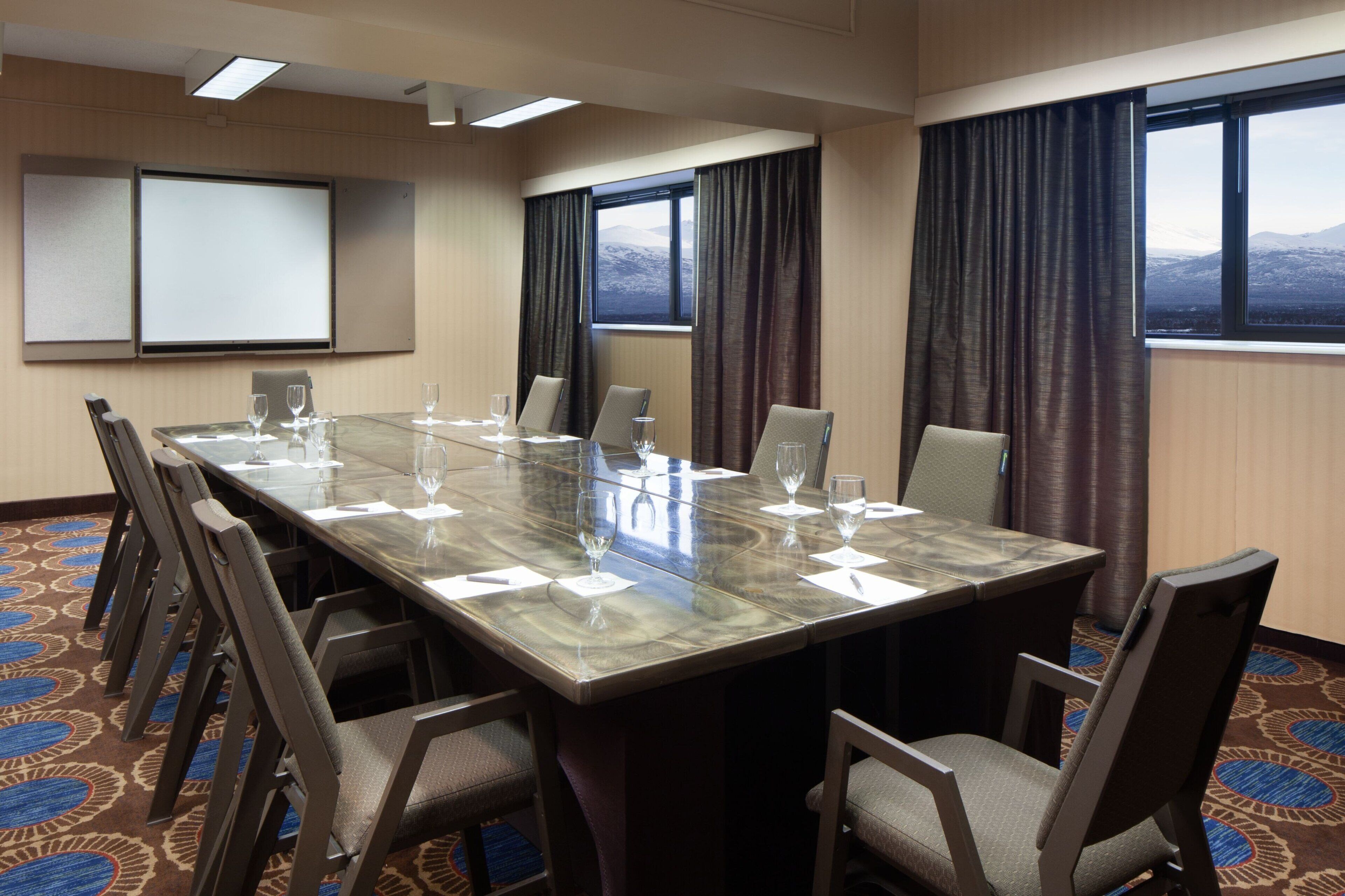 A conference room with a large table, chairs, a projector screen, and windows with curtains.