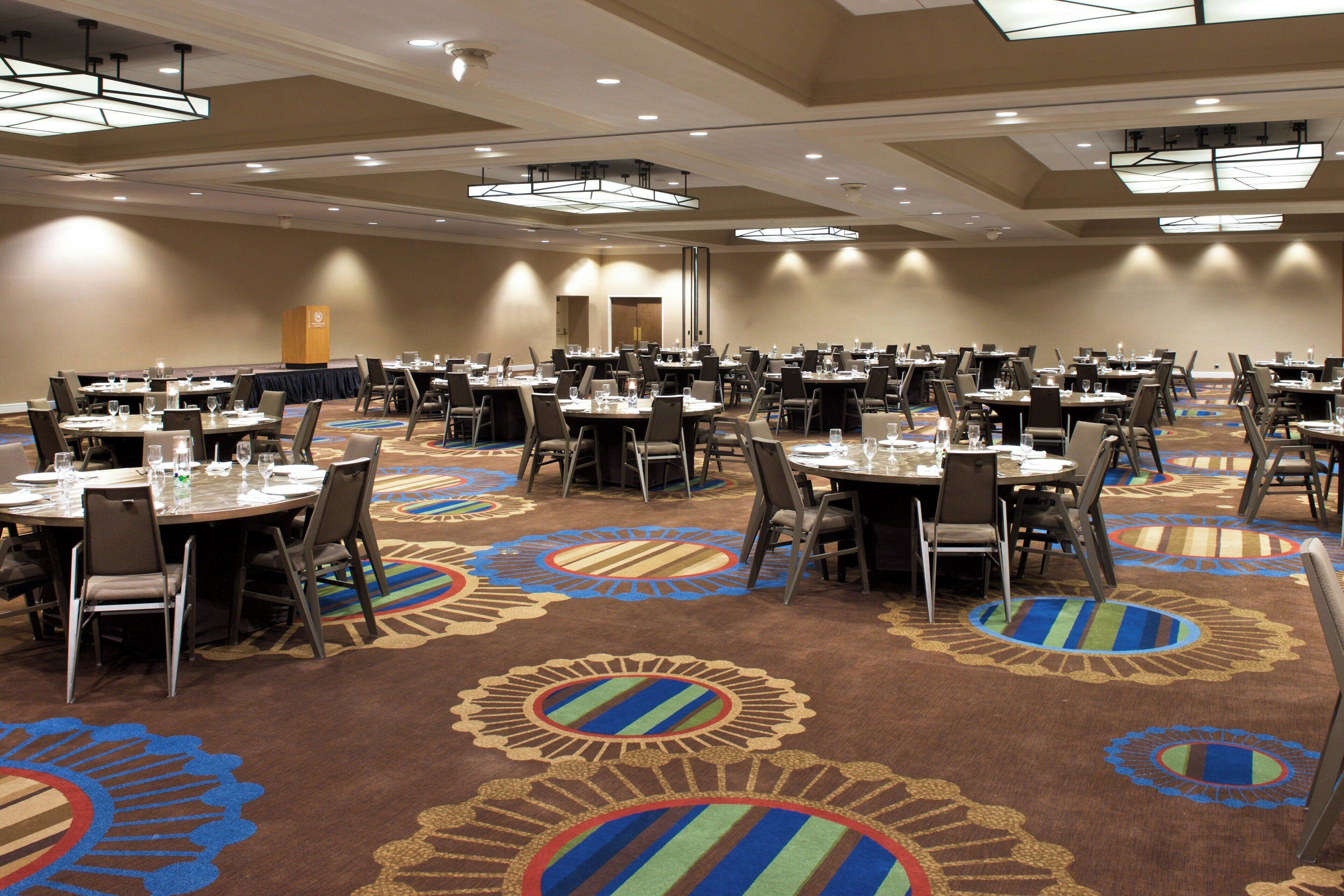 Une grande salle de conférence dotée de nombreuses tables et chaises, d’un sol recouvert de moquette orné de motifs circulaires et d’un éclairage au plafond.