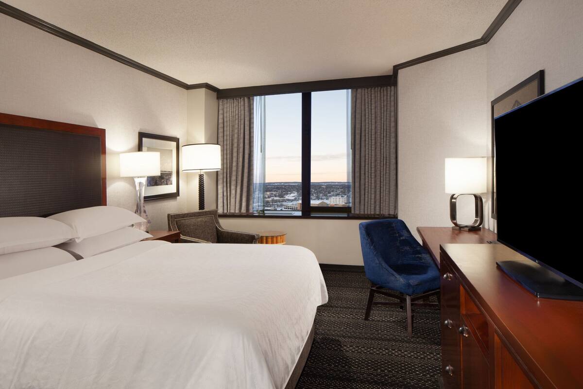 Quarto de hotel com uma cama grande, uma escrivaninha com televisão de tela plana, uma cadeira e uma janela com vista para a cidade.