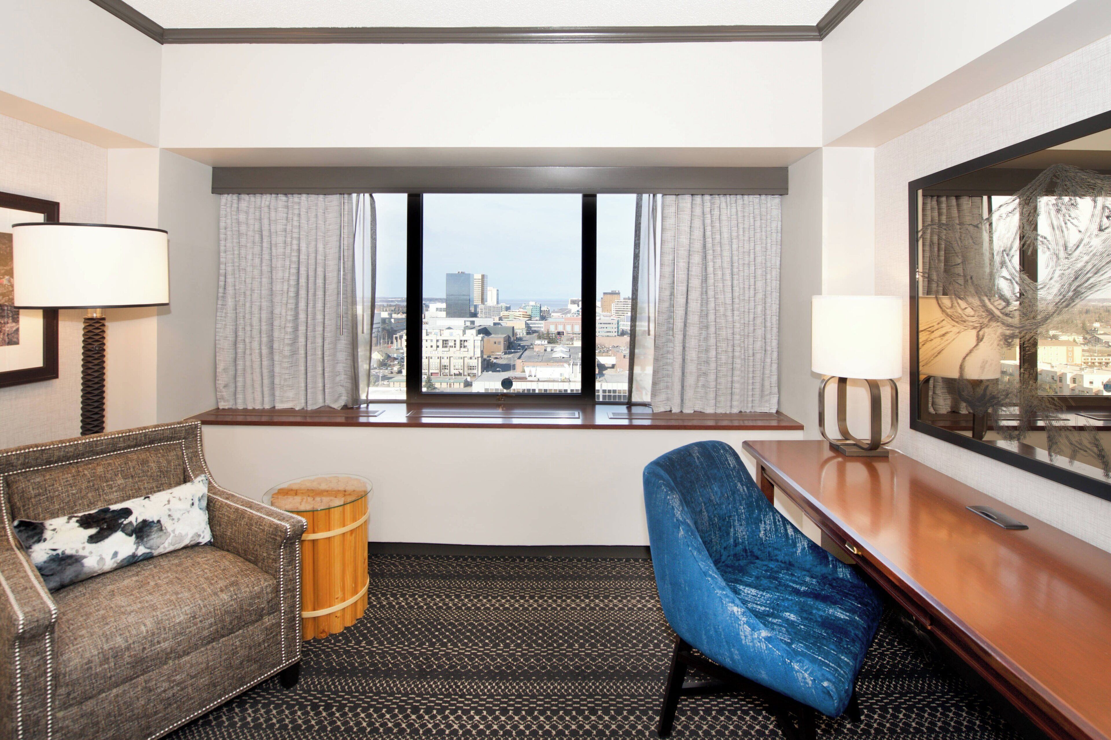 Une chambre d’hôtel avec vue sur la ville, comprenant un canapé, un fauteuil, un tabouret en bois et un bureau avec une lampe.