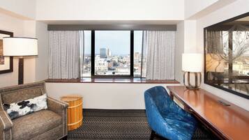 A hotel room with a city view, a sofa, a chair, a wooden stool, and a desk with a lamp.