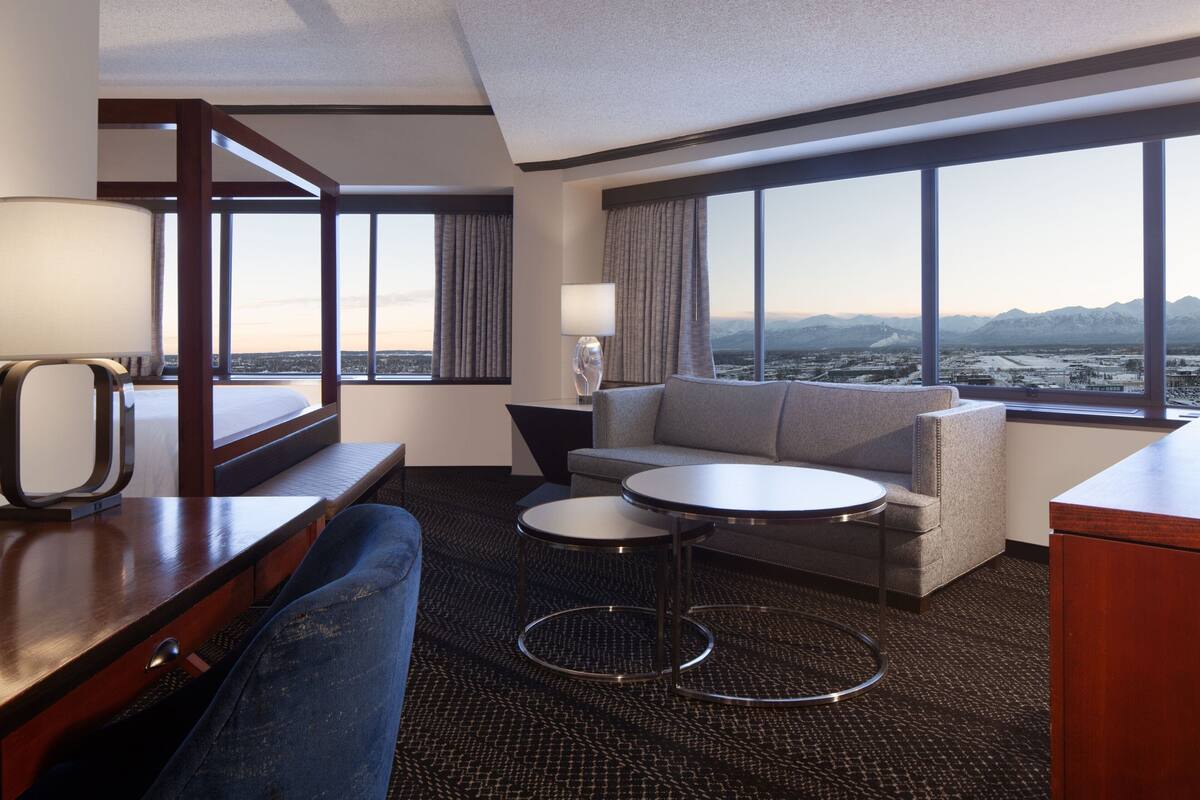 Quarto de hotel com cama, sofá, escrivaninha, luminária, mesa redonda e amplas janelas com vista para a cidade e montanhas.