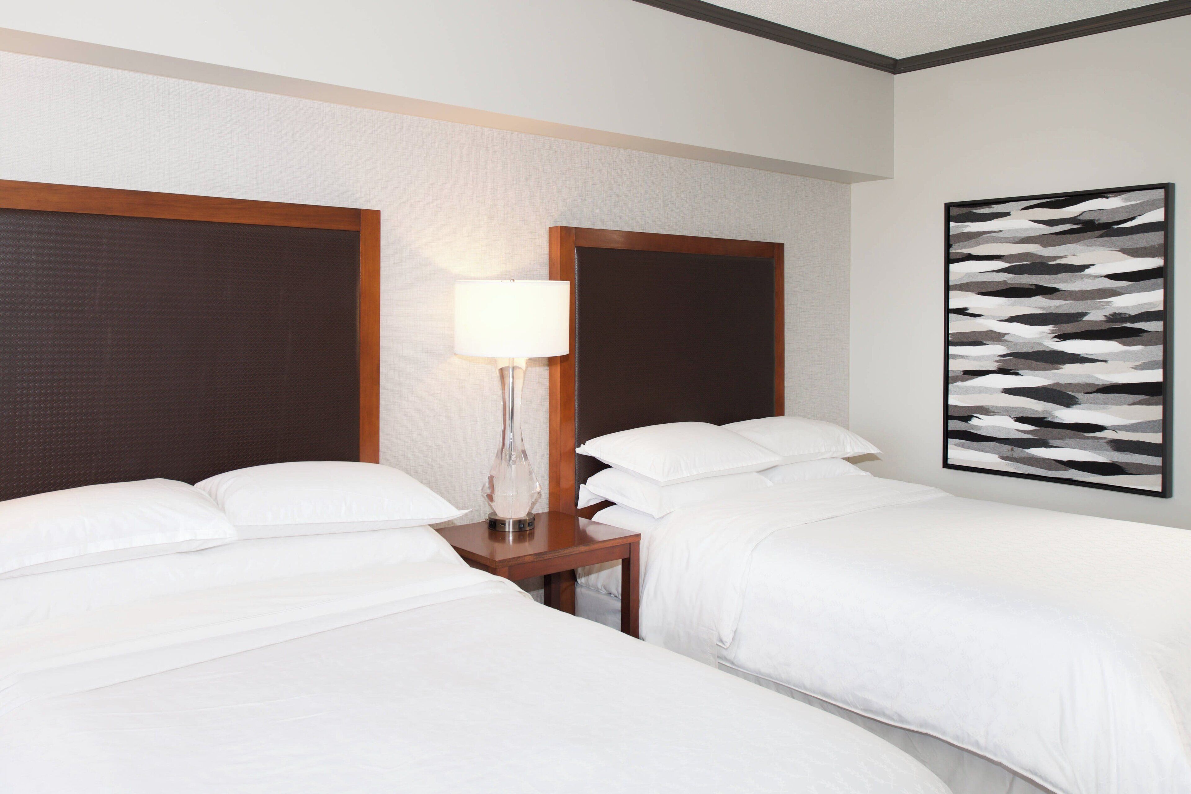 Habitación de hotel con dos camas, una mesita de noche con lámpara y una obra de arte abstracto colgada en la pared.