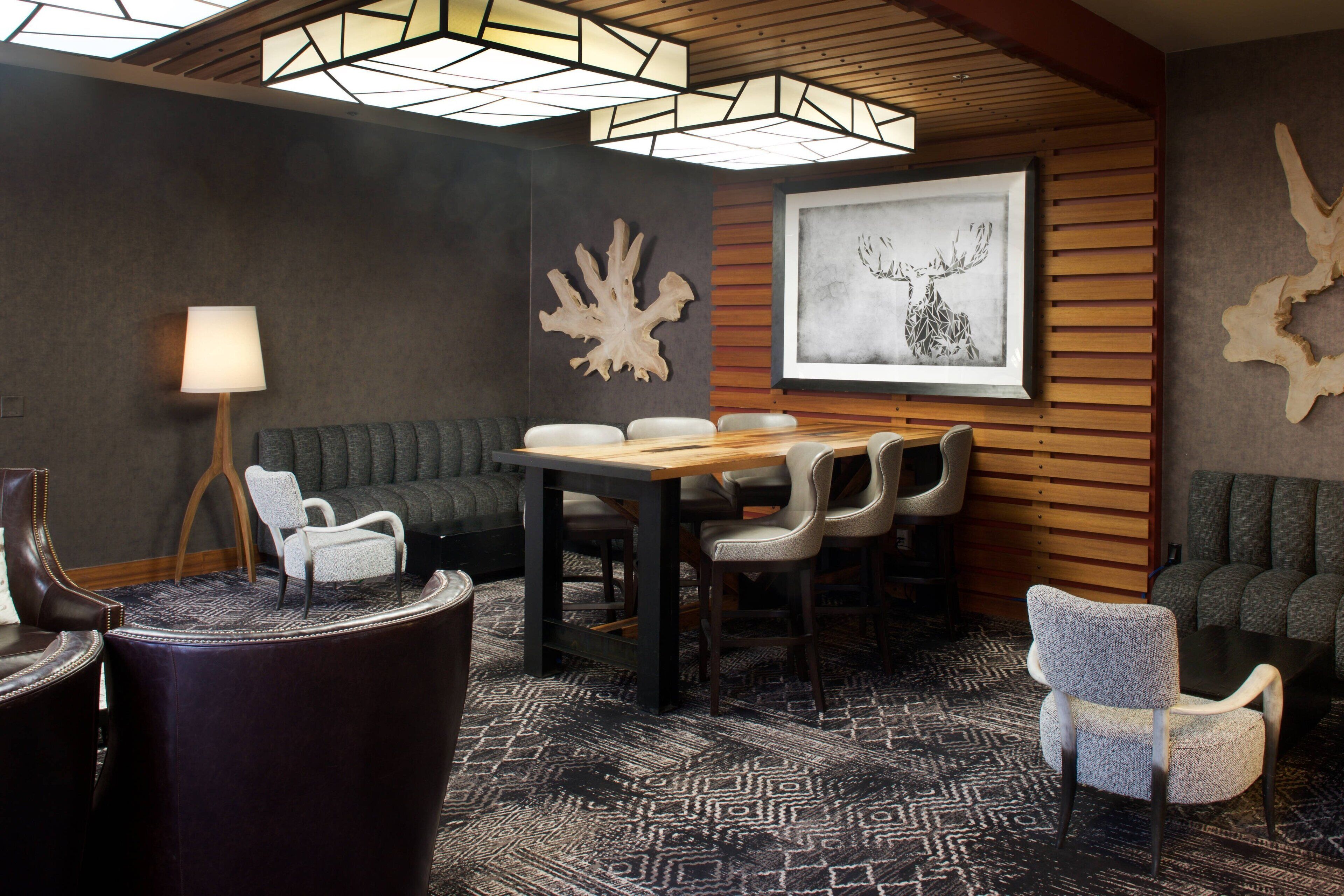 A modern lounge area with a wooden table, leather chairs, a patterned carpet, and a ceiling with geometric light fixtures.