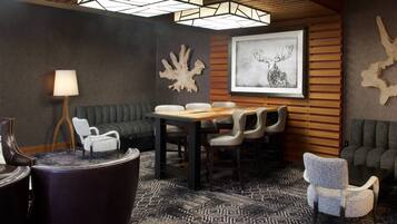 A modern lounge area with a wooden table, leather chairs, a patterned carpet, and a ceiling with geometric light fixtures.