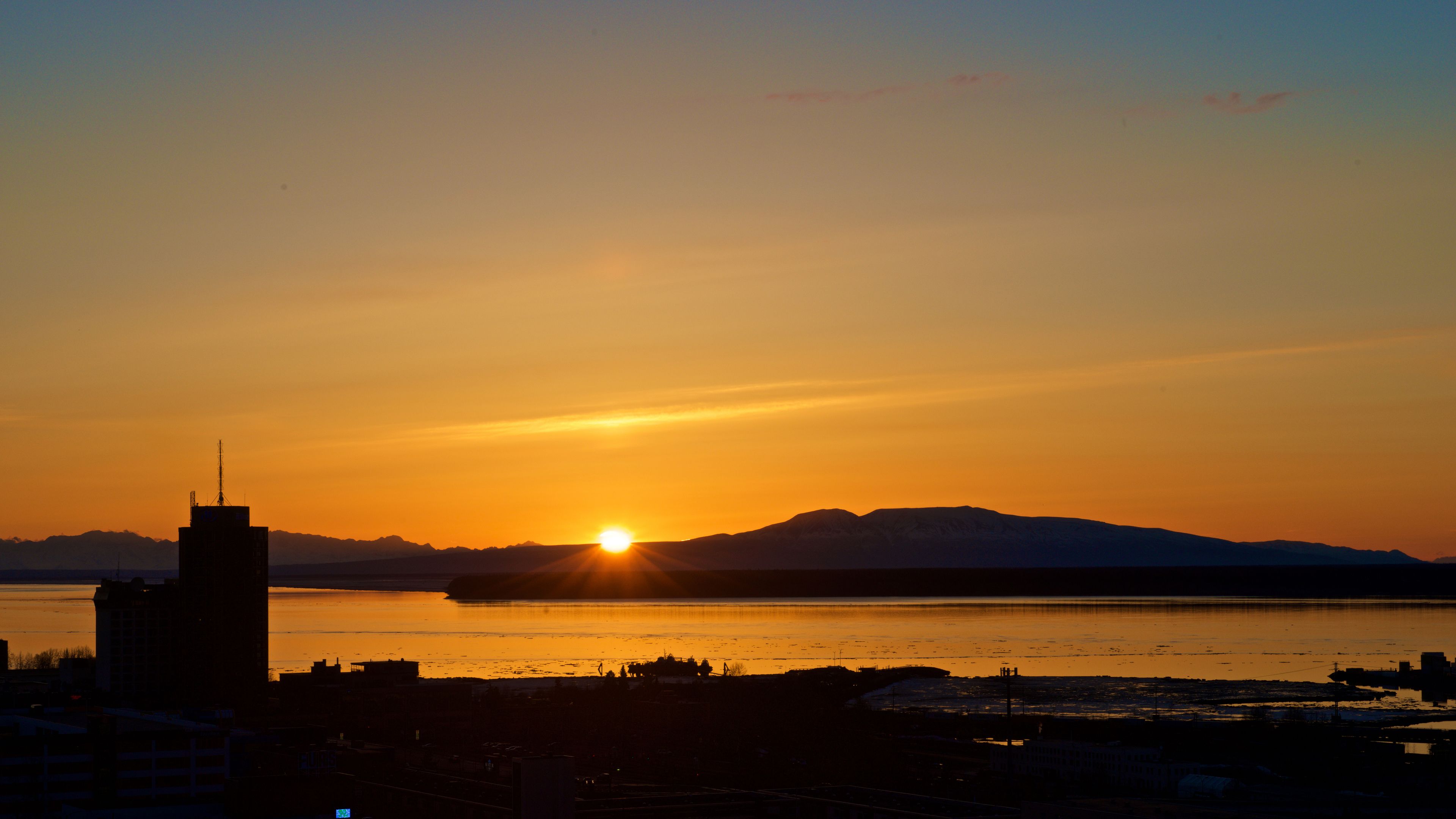 A sunset over a body of water with a silhouette of a cityscape and mountains in the background.