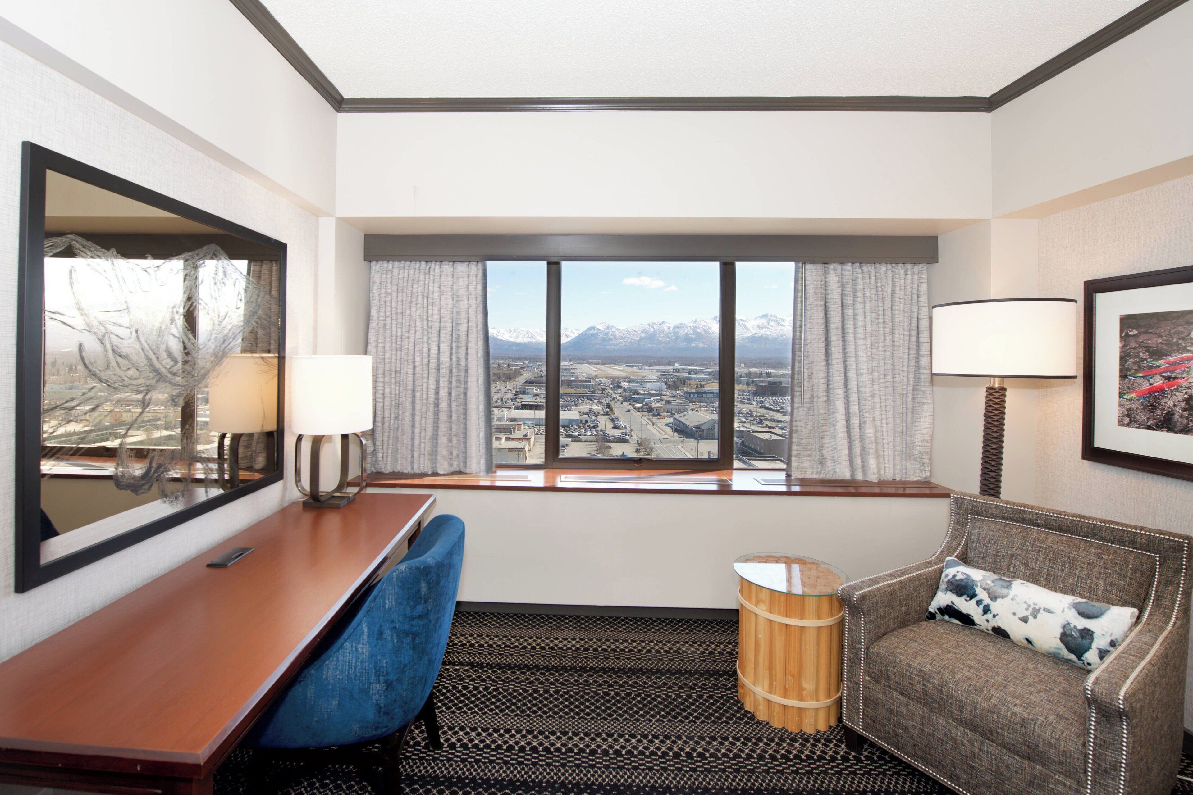 A hotel room with a desk, chair, and a view of mountains through large windows.