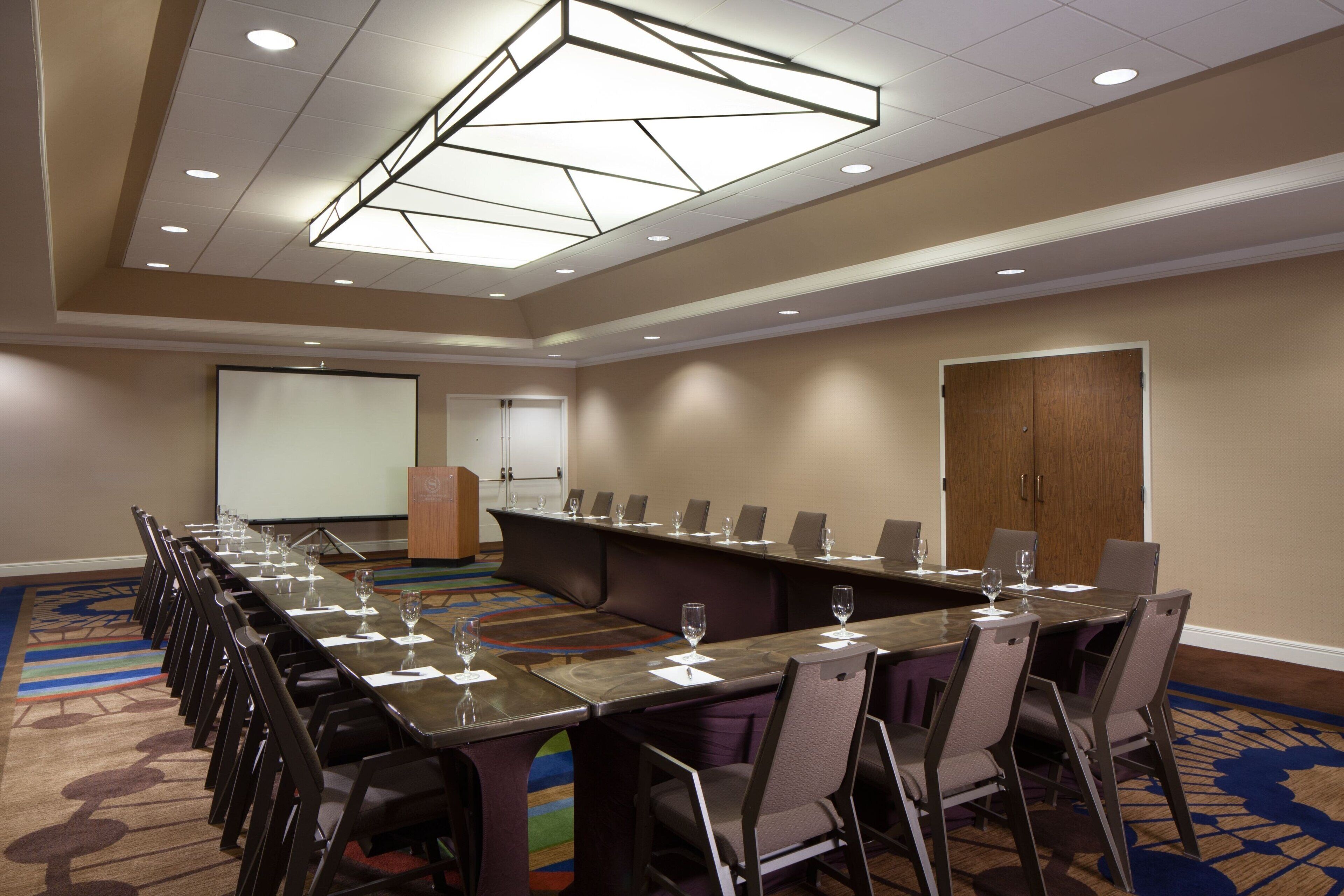 A conference room with a long table, chairs, a projector screen, and a carpeted floor.