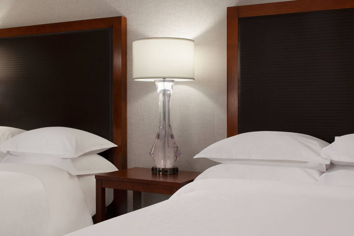 Quarto de hotel com cama, almofadas brancas, mesa de cabeceira e abajur.