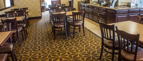 Un restaurante con alfombra estampada, mesas y sillas de madera, una zona de bar y un televisor en la pared.