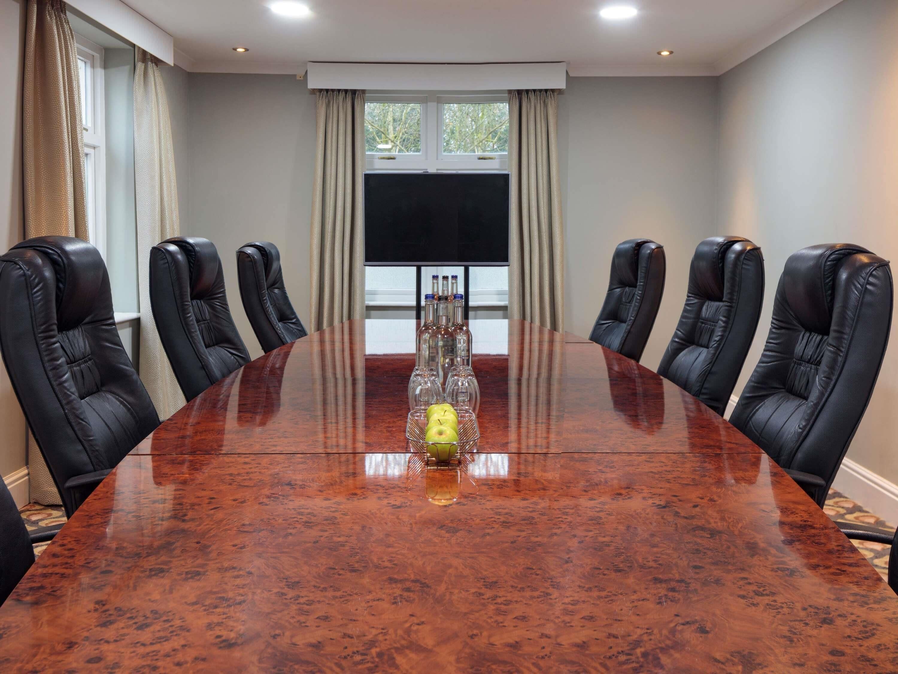 Une salle de conférence dotée d’une grande table en bois, de fauteuils en cuir noir, d’un téléviseur à écran plat et d’une fenêtre avec des rideaux.