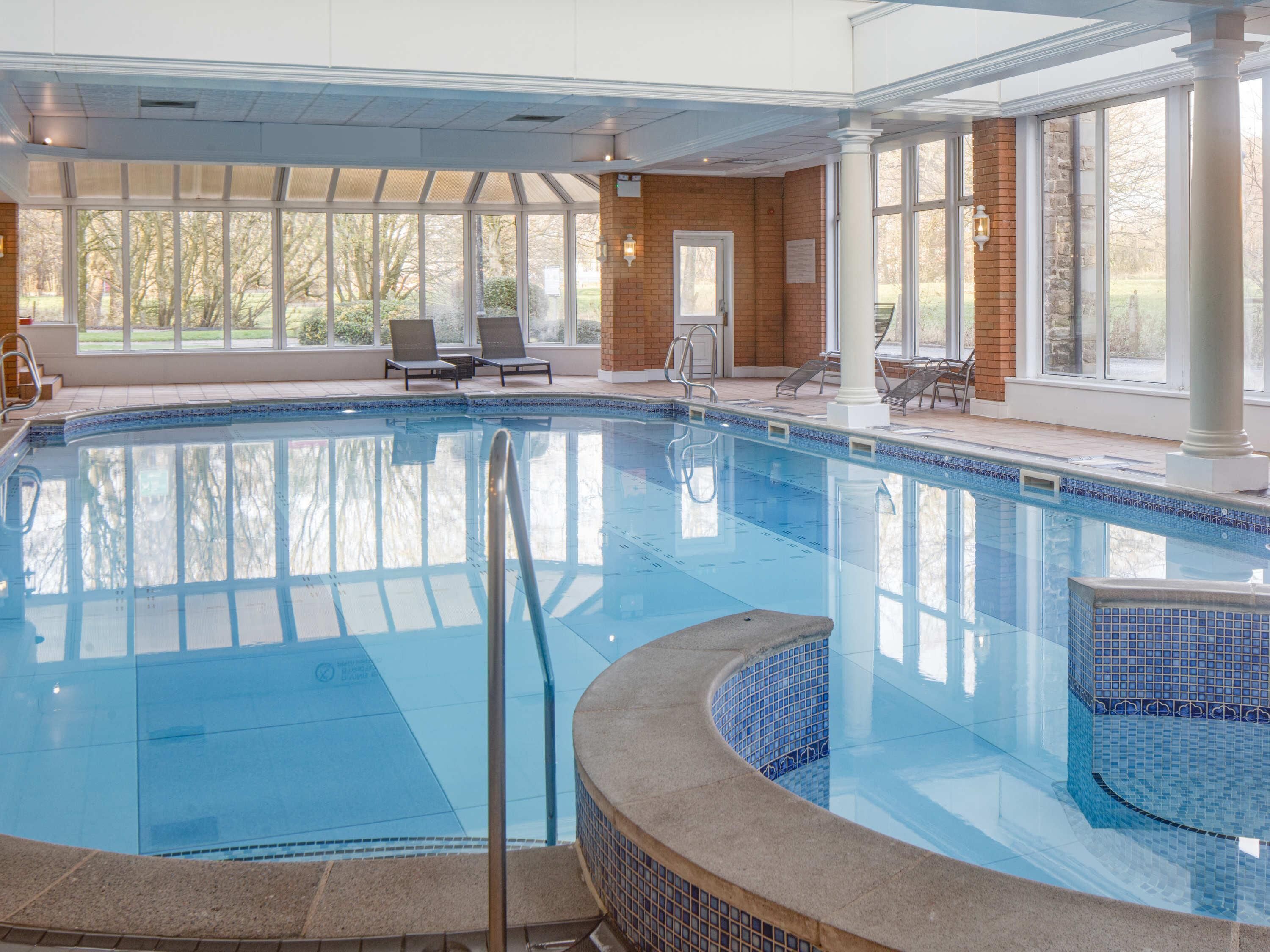 An indoor swimming pool with clear blue water, a tiled edge, and a sunlit area with windows and chairs.