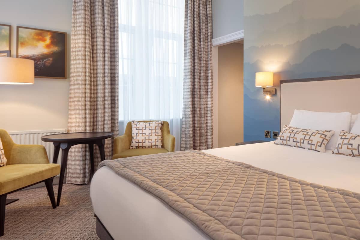 A hotel room with a large bed, two armchairs, a small table, and a view of the outdoors through sheer curtains.