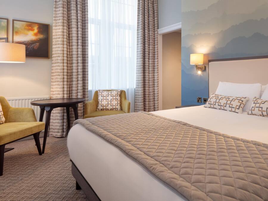 A hotel room with a large bed, two armchairs, a small table, and a view of the outdoors through sheer curtains.