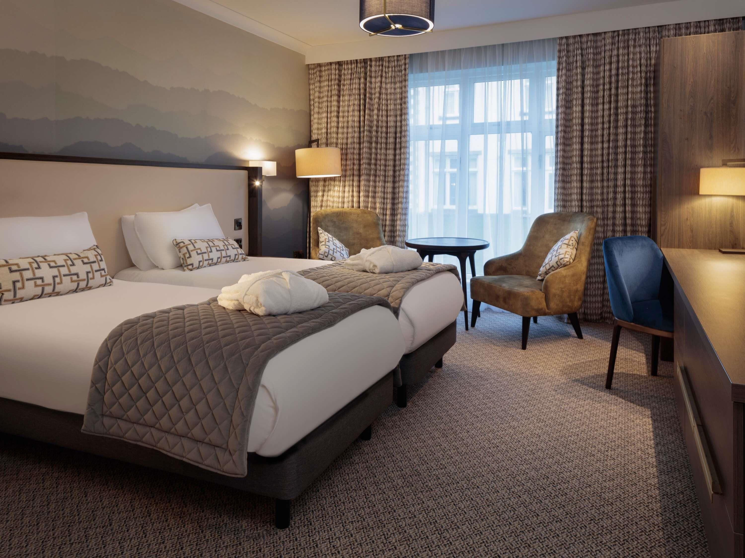 A hotel room with a large bed, two armchairs, a small table, and a view of the outdoors through a window.