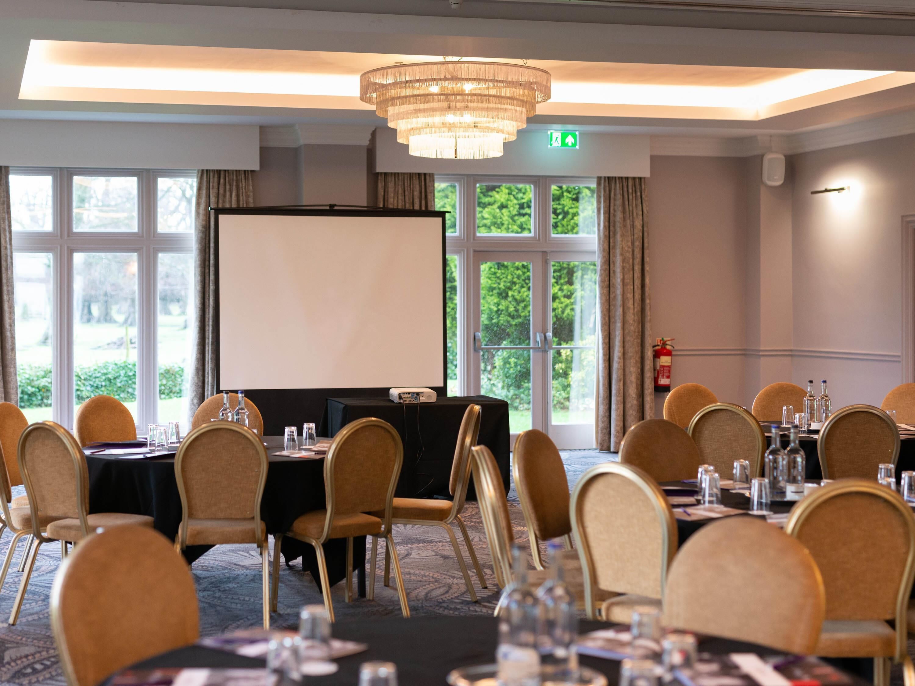 A conference room with tables, chairs, a projector screen, and a large window with a view of greenery.