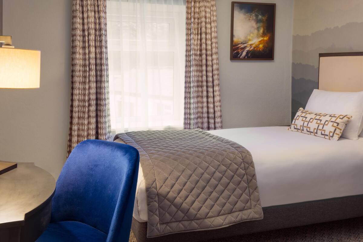 A hotel room with a bed, a chair, a lamp, and a painting on the wall.