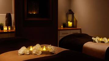 Una habitación tenuemente iluminada con dos camas, una mesita con velas, un espejo y un jarrón decorativo.