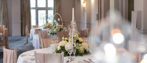 Un salón de banquetes con mesas redondas dispuestas para un evento formal, con centros de mesa florales y una elegante vajilla.
