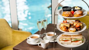 Ein mehrstufiges Desserttablett mit Gebäck und Sandwiches, zwei Kaffeetassen und zwei Sektgläser auf einem Tisch am Pool.