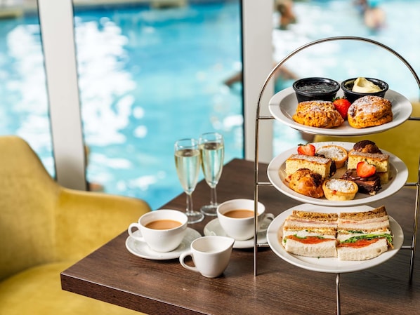 Ein mehrstufiges Desserttablett mit Gebäck und Sandwiches, zwei Kaffeetassen und zwei Sektgläser auf einem Tisch am Pool.