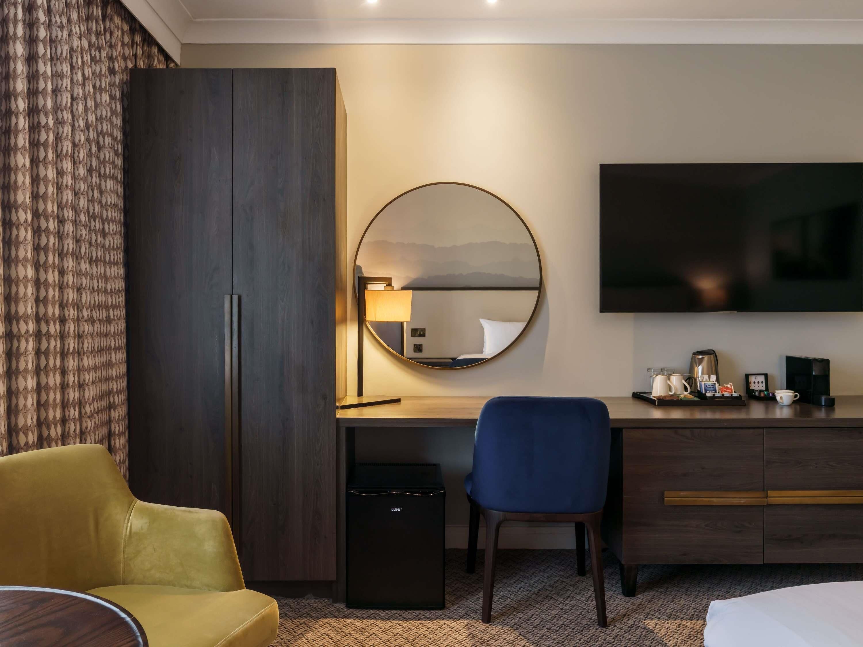 Une chambre d’hôtel comprenant un lit, un bureau avec une chaise, une armoire, un miroir, une télévision et une machine à café.