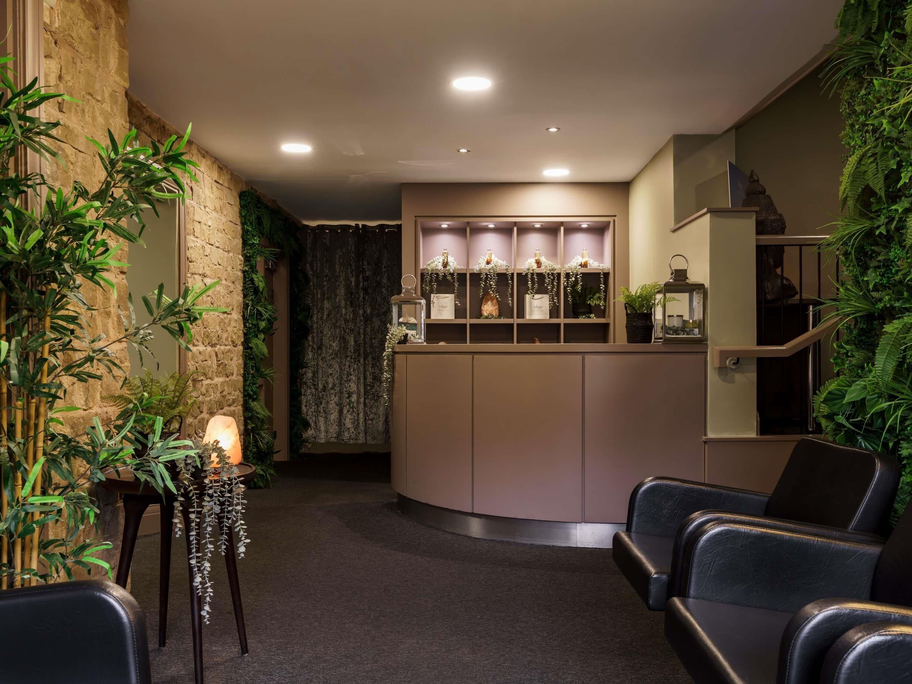 A reception area with a curved counter, leather chairs, a small table with a lamp, and a wall with green plants.