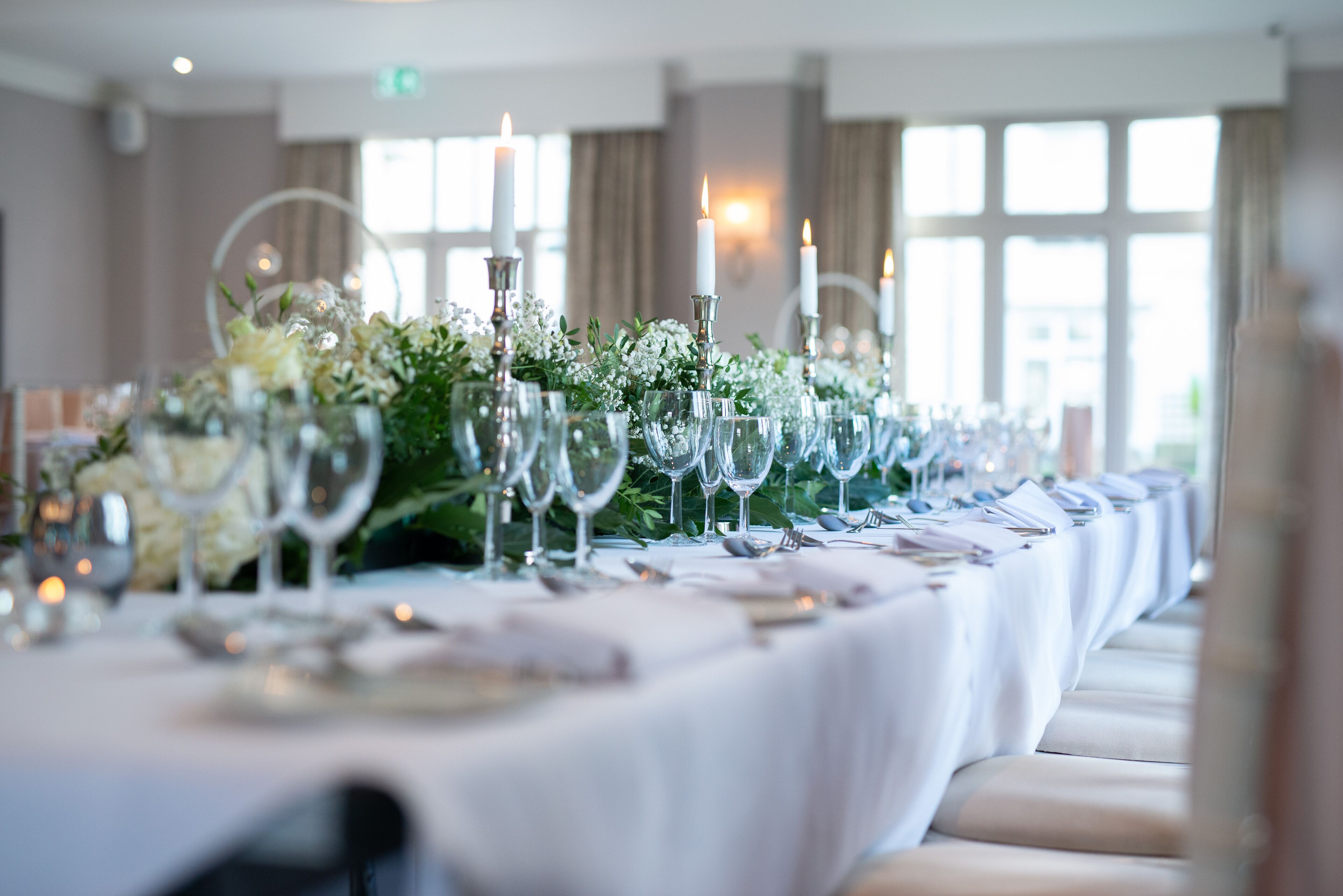 A long banquet table set with white tablecloths, glassware, and candles, in a room with large windows and curtains.