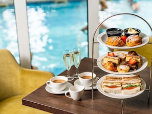 Suporte de sobremesa de três andares com doces e sanduíches, duas xícaras de café e um copo de água em uma mesa perto de uma piscina.