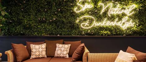 A modern living room with a brown sofa and a wicker chair, a green wall with a neon sign reading "Spa Nature Dunkelh".