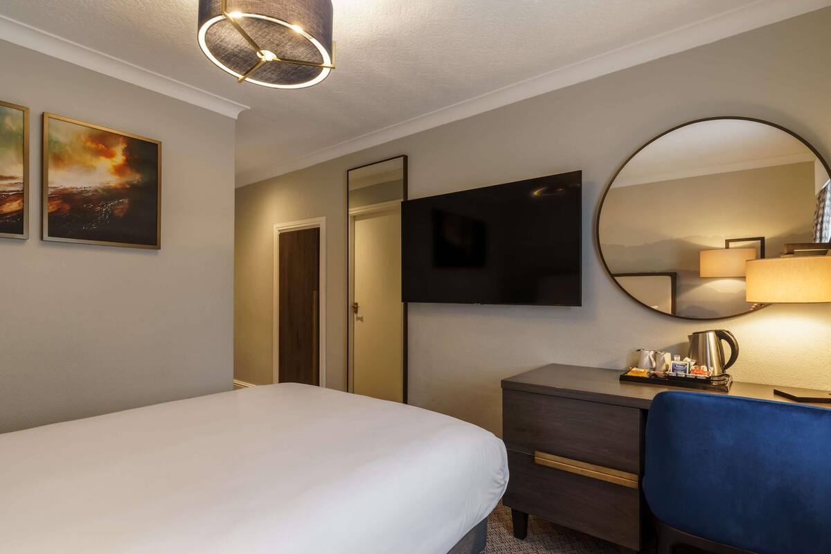 A hotel room with a bed, a desk, a chair, a television, a mirror, and a lamp.