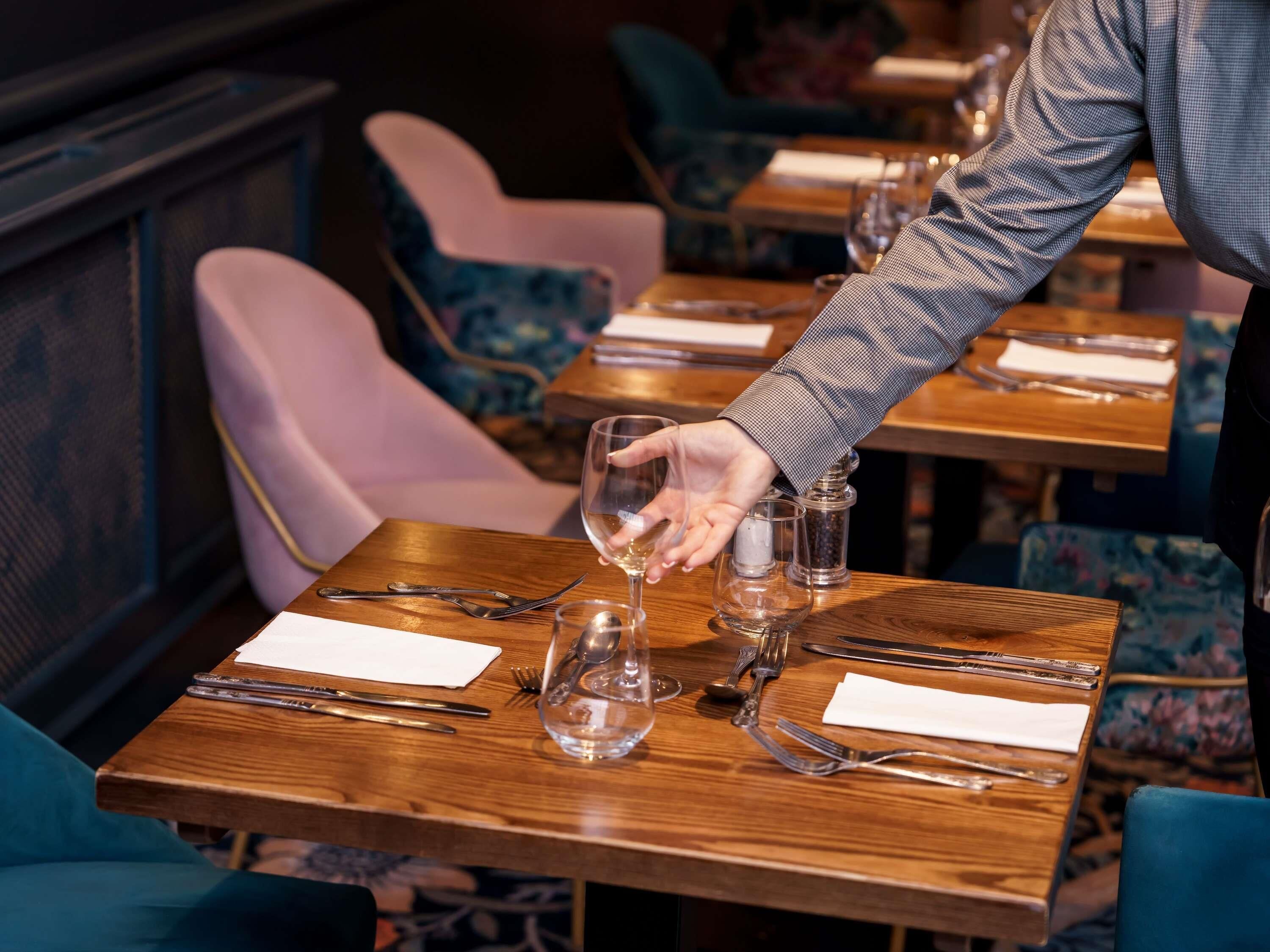 A restaurant setting with wooden tables, chairs, and a person pouring a drink.