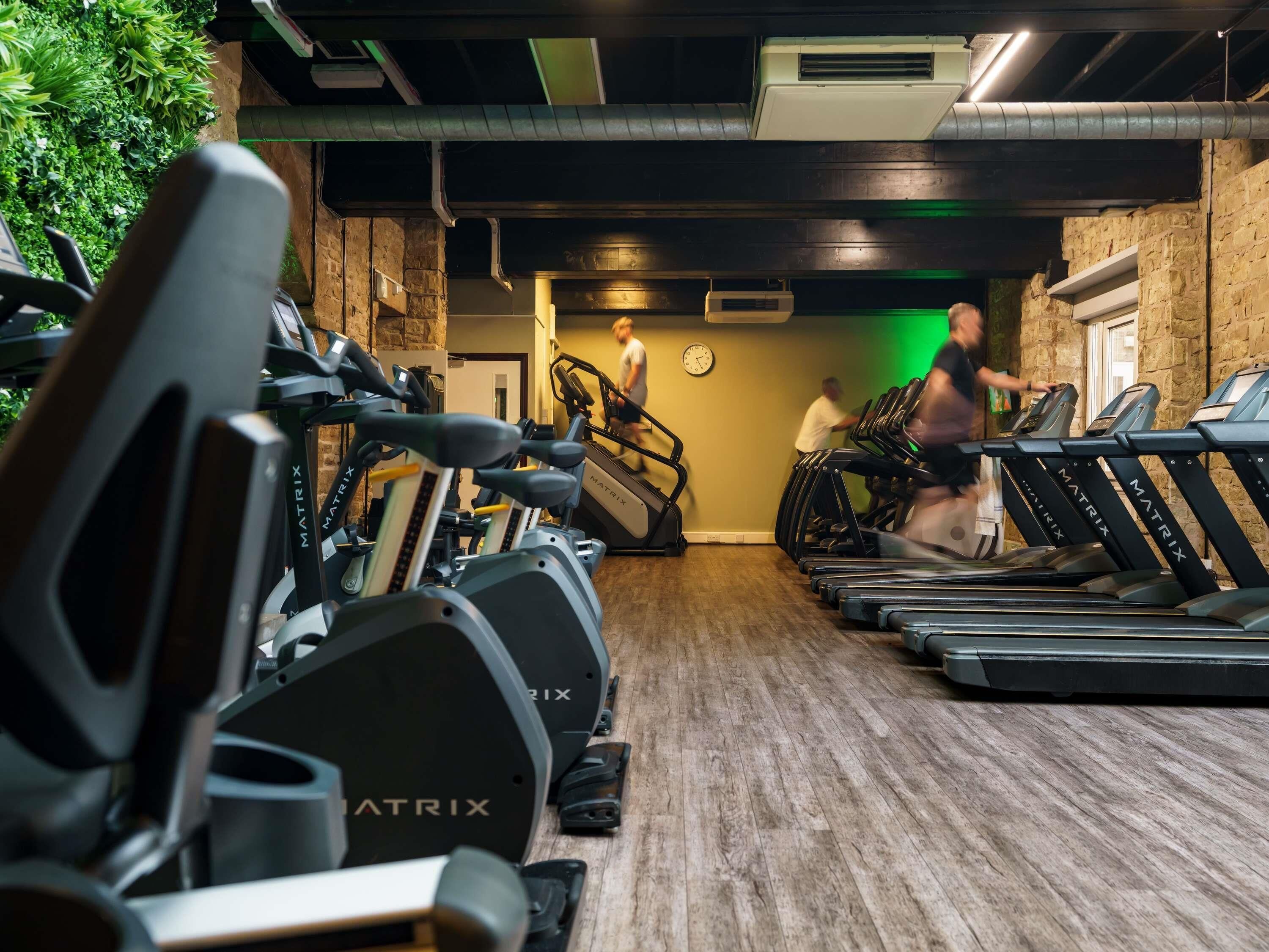 A gym with multiple treadmills and stationary bikes, a clock on the wall, and a green-lit ceiling.
