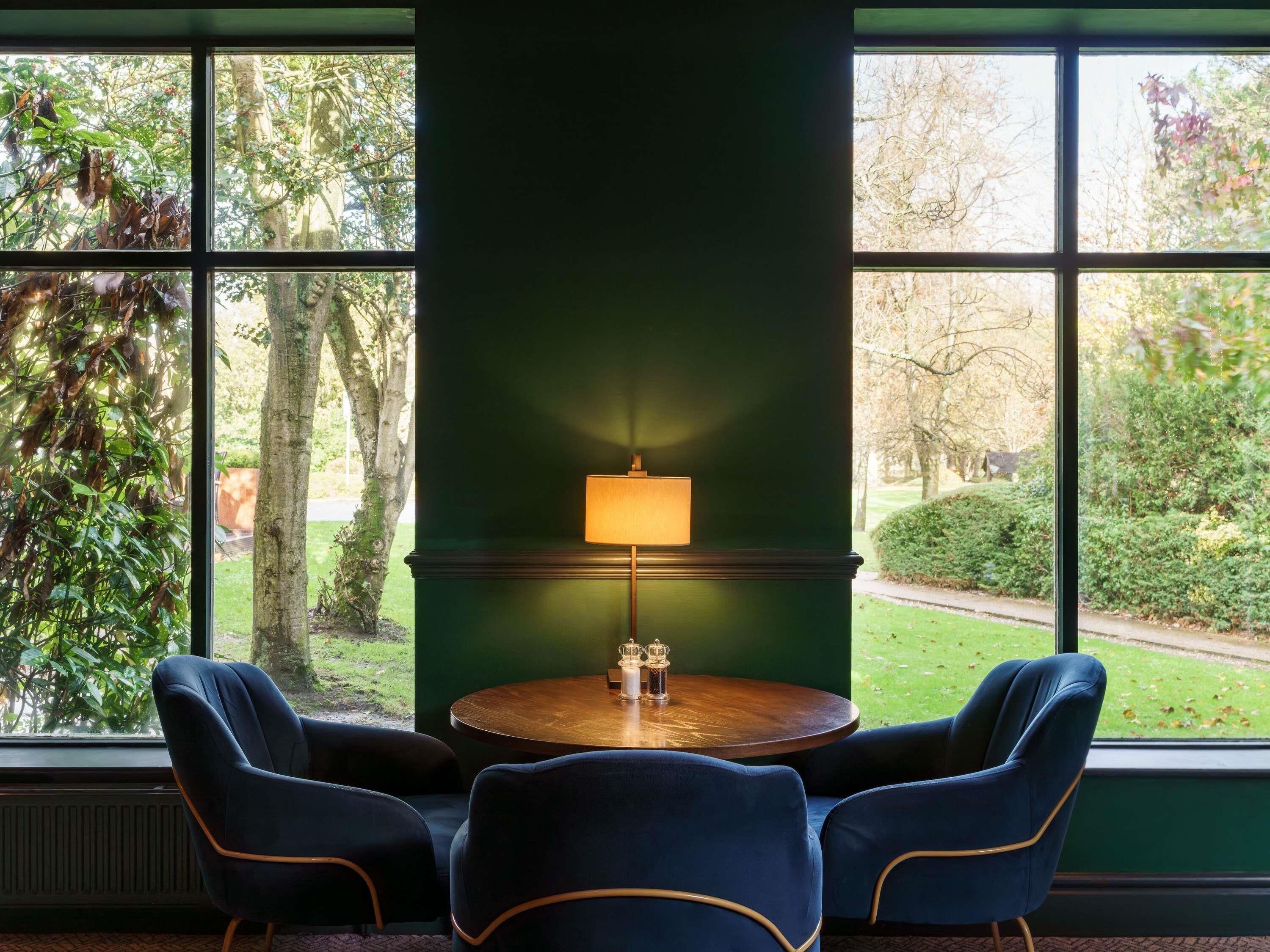 Una habitación con una mesa redonda de madera, dos sillas azules, una pared verde y una ventana con vista a un jardín.