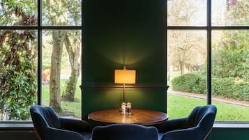 Una habitación con una mesa redonda de madera, dos sillas azules, una pared verde y una ventana con vista a un jardín.