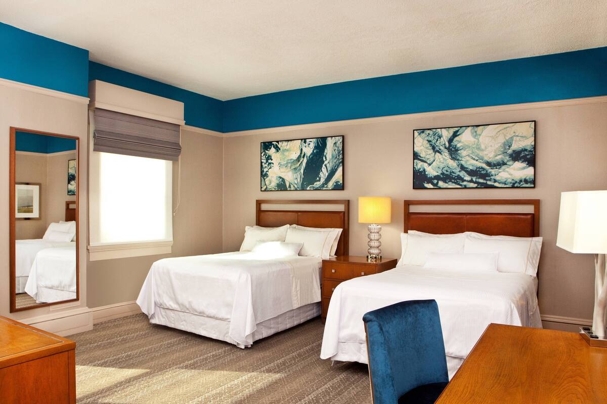 A hotel room with two beds, a wooden desk, a chair, a nightstand with a lamp, and artwork on the walls.