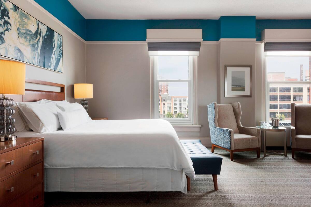 A hotel room with a large bed, two chairs, a small table, and a window with a city view.