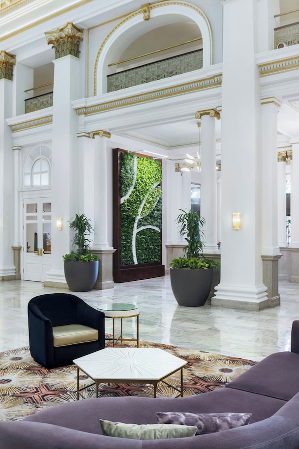 Eine geräumige Lobby mit hohen Decken, großen Säulen und einer zentralen Tür mit einer grünen Wand. Es gibt Kübelpflanzen, einen runden Tisch und eine Couch mit Kissen.