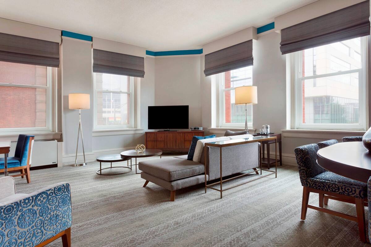 A modern hotel room with a sofa, armchair, dining table, and a flat-screen TV.