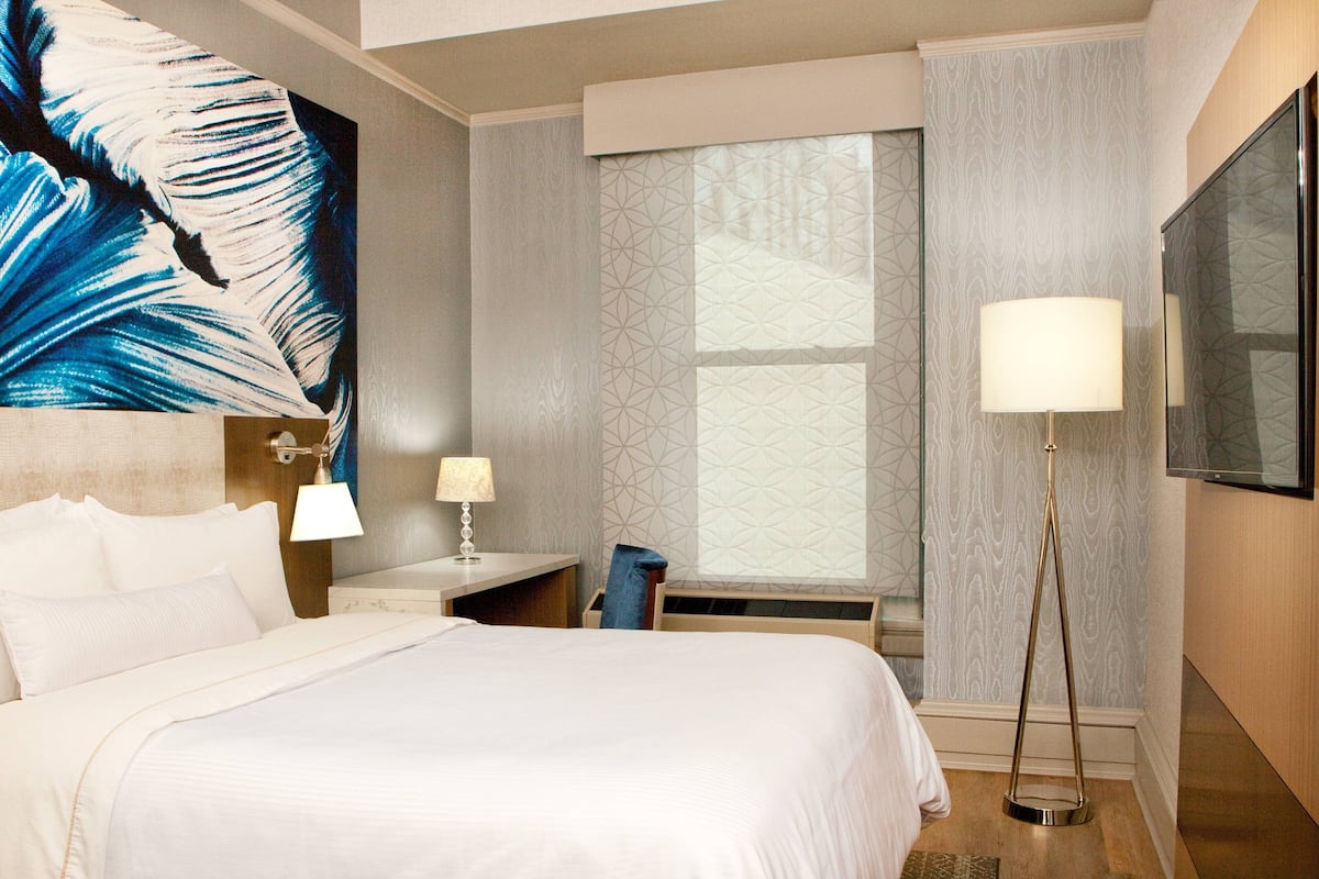 A hotel room with a large bed, a desk with a lamp, a chair, and a large window with patterned curtains.