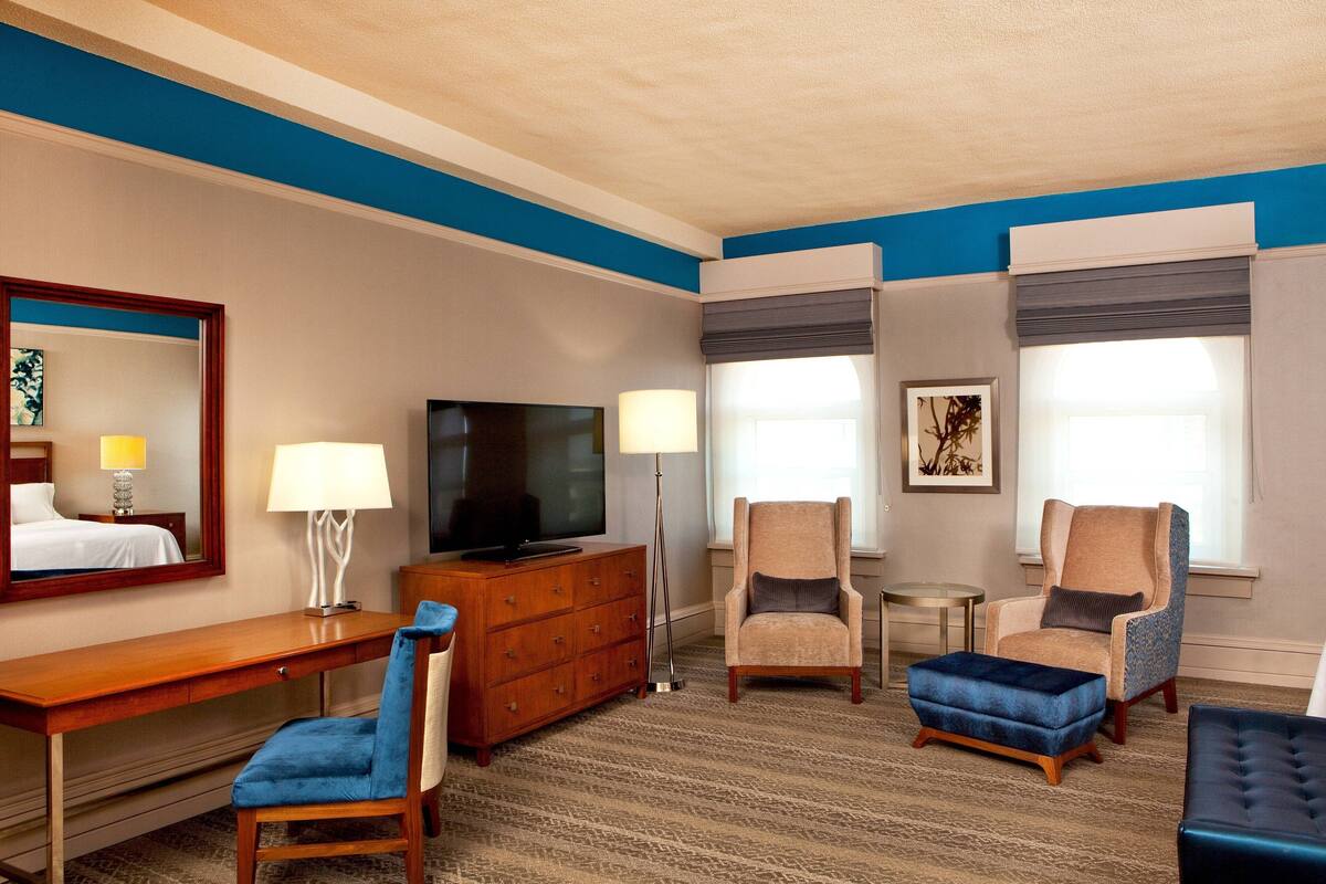 A hotel room with a wooden desk, two armchairs, a blue ottoman, a television, and a lamp.