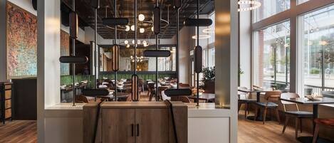 A modern restaurant interior with exposed ceilings, hanging pendant lights, and a view of the dining area through large windows.