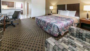 Habitación de hotel con una cama grande, un escritorio con silla, una kitchenette pequeña y un baño visible a través de una puerta abierta.