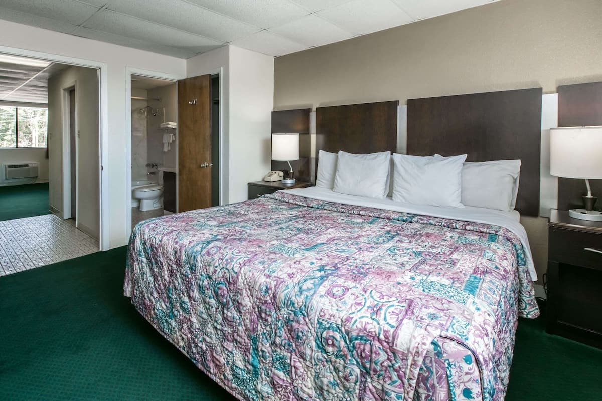 A hotel room with a large bed, a nightstand with a lamp, a wooden headboard, and a bathroom visible through an open door.
