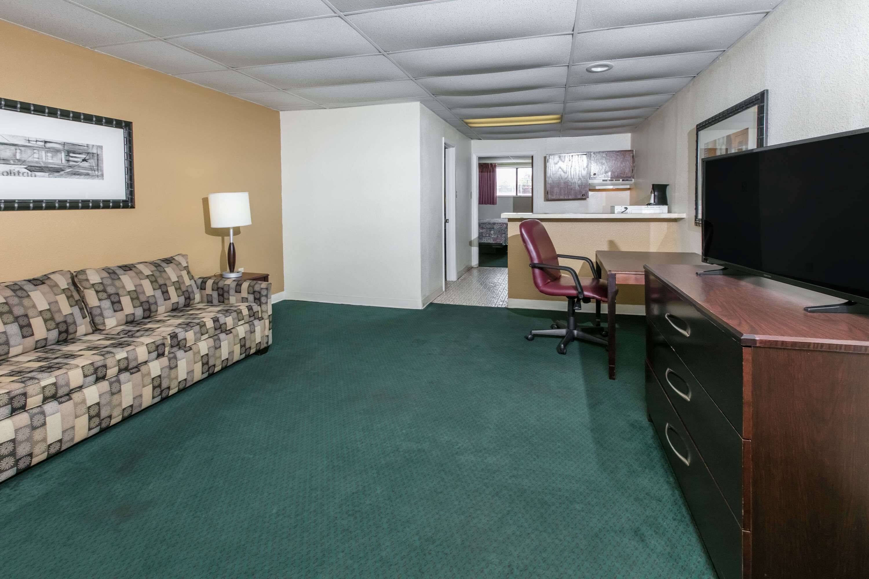 Habitación de hotel con alfombra verde, sofá, escritorio con televisor, una silla y una zona de cocina al fondo.