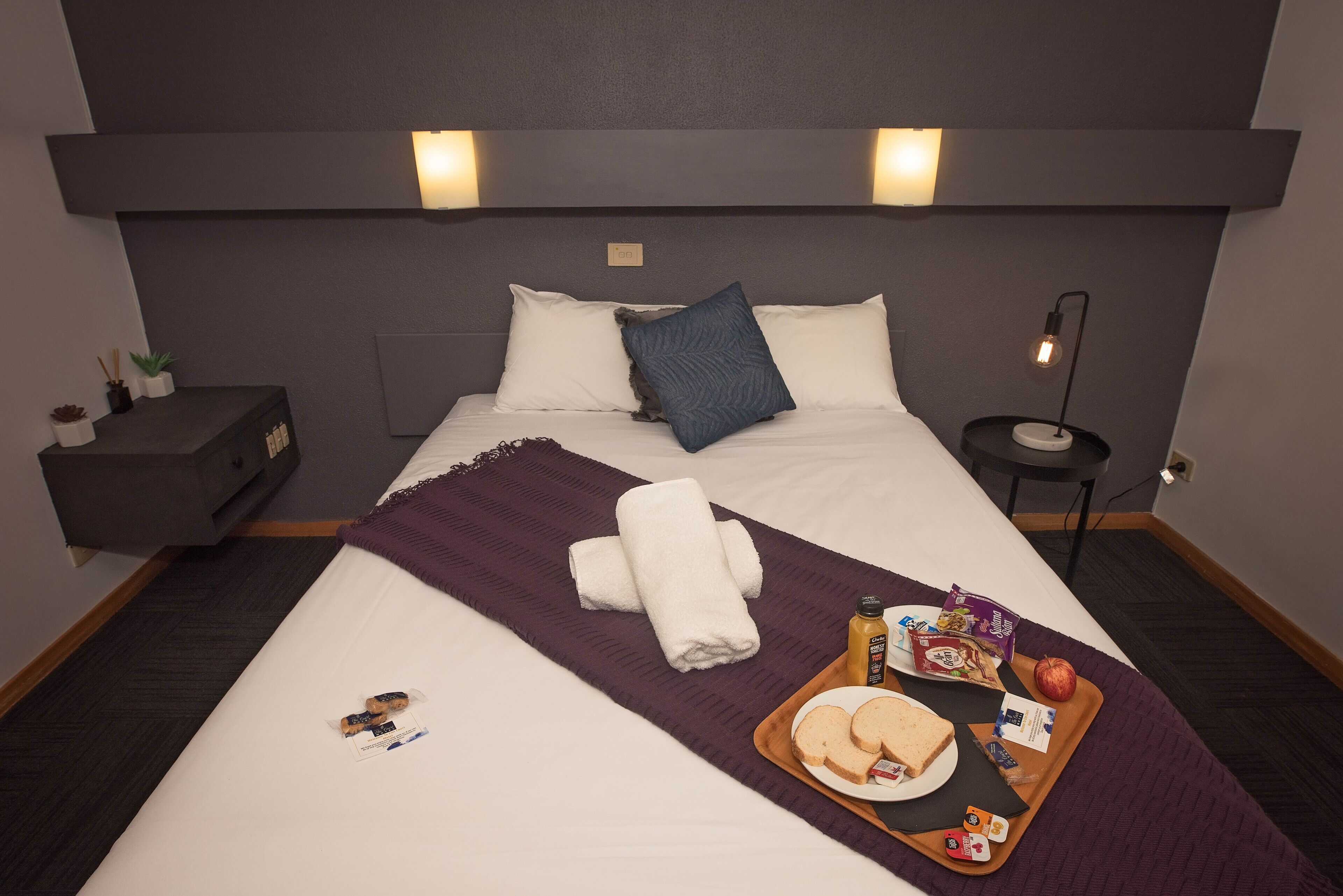 Cama bem arrumada com manta roxa, bandeja com itens de café da manhã e mesa de cabeceira com abajur.